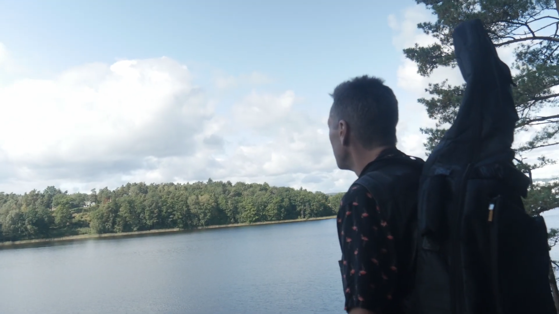 Stillbild ur Lycklig Och Fri. Person med gitarrfodral blickar ut över en sjö